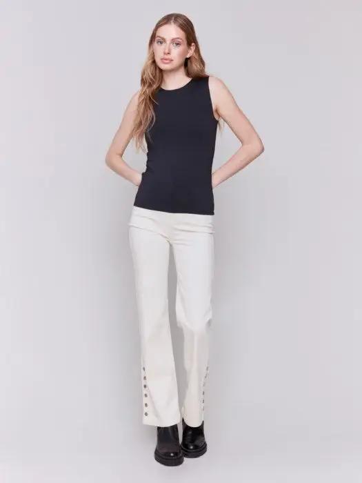 A person with long curly hair, wearing a Charlie B Super Stretch Sleeveless Top, blue jeans, and black ankle boots, stands posing against a plain background.