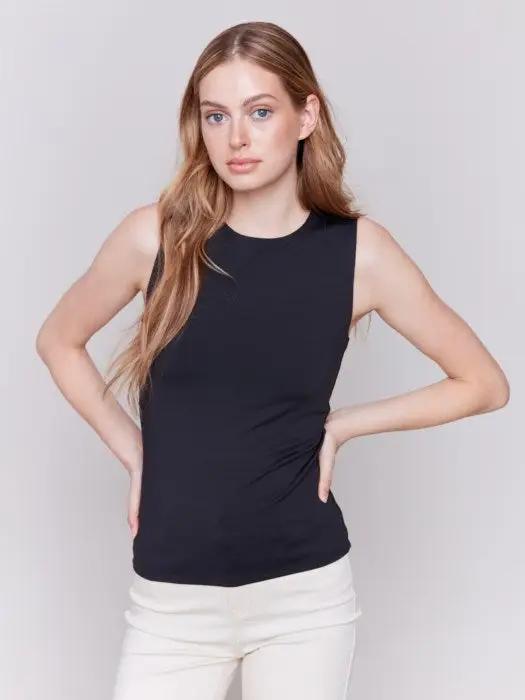 A person with long curly hair, wearing a Charlie B Super Stretch Sleeveless Top, blue jeans, and black ankle boots, stands posing against a plain background.