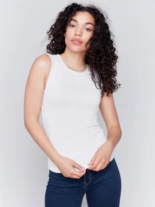 A person with long curly hair, wearing a Charlie B Super Stretch Sleeveless Top, blue jeans, and black ankle boots, stands posing against a plain background.
