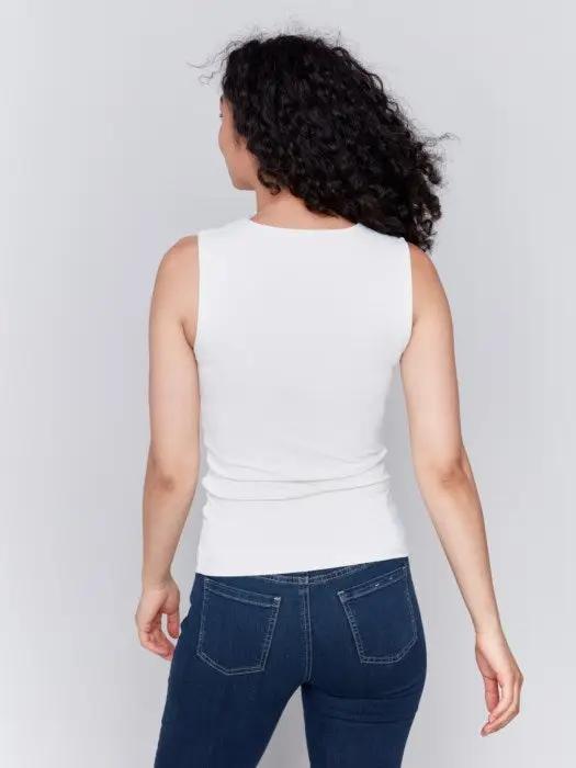 A person with long curly hair, wearing a Charlie B Super Stretch Sleeveless Top, blue jeans, and black ankle boots, stands posing against a plain background.