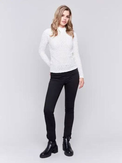 A woman with wavy blonde hair wearing a stylish Charlie B Basic Mock Neck Sweater and black pants stands against a light gray background.