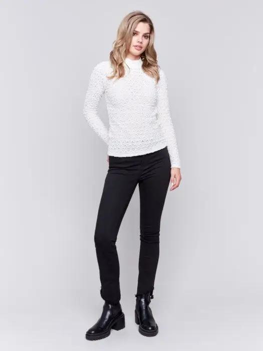 A woman with wavy blonde hair wearing a stylish Charlie B Basic Mock Neck Sweater and black pants stands against a light gray background.