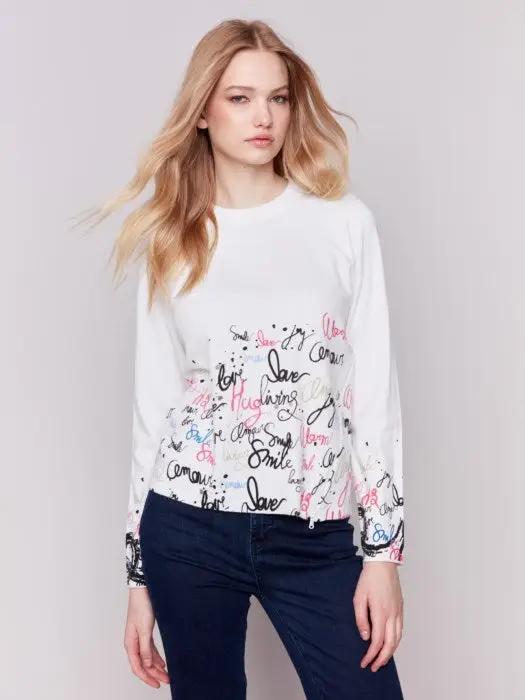 A woman is wearing a trendy Charlie B Printed Sweater featuring colorful words like "love" and "smile" along with a diagonal zipper, standing against a plain background.