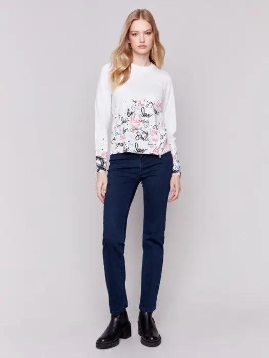 A woman is wearing a trendy Charlie B Printed Sweater featuring colorful words like "love" and "smile" along with a diagonal zipper, standing against a plain background.