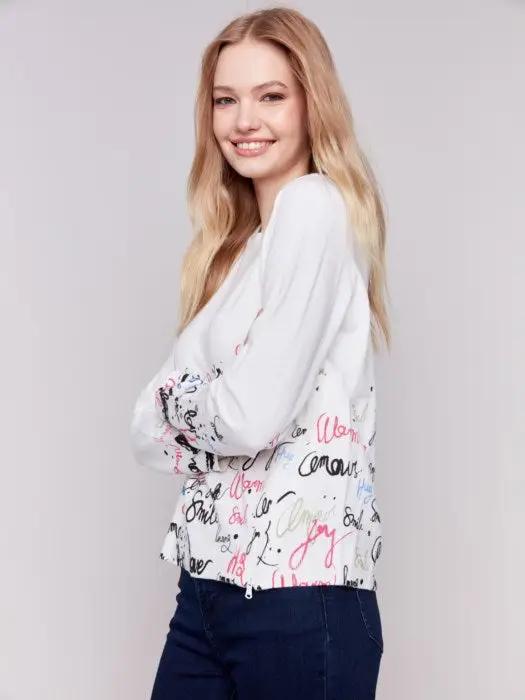 A woman is wearing a trendy Charlie B Printed Sweater featuring colorful words like "love" and "smile" along with a diagonal zipper, standing against a plain background.