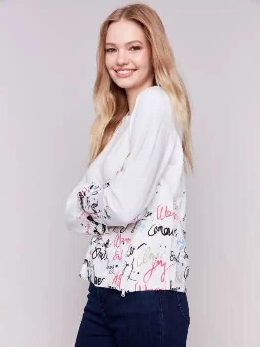 A woman is wearing a trendy Charlie B Printed Sweater featuring colorful words like "love" and "smile" along with a diagonal zipper, standing against a plain background.