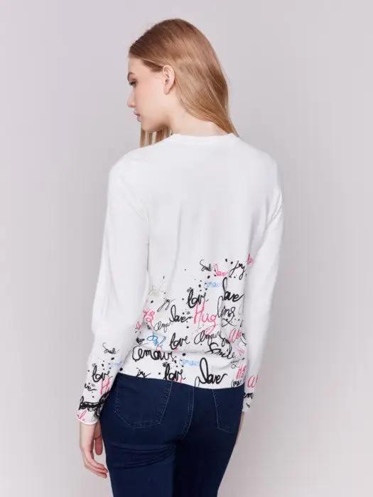 A woman is wearing a trendy Charlie B Printed Sweater featuring colorful words like "love" and "smile" along with a diagonal zipper, standing against a plain background.
