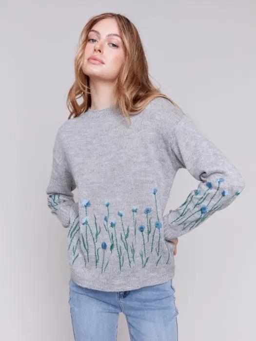 A woman with long hair is wearing the Charlie B Plush Knit Embroidered Flower Sweater featuring gray fabric adorned with blue and green floral details, paired with light blue jeans, creating a look that exudes cozy warmth.