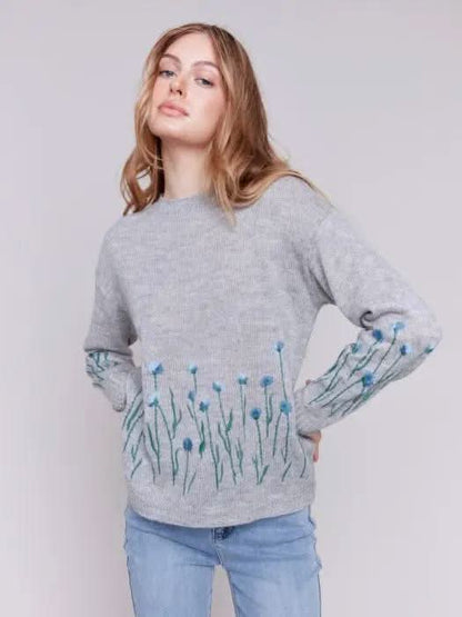A woman with long hair is wearing the Charlie B Plush Knit Embroidered Flower Sweater featuring gray fabric adorned with blue and green floral details, paired with light blue jeans, creating a look that exudes cozy warmth.