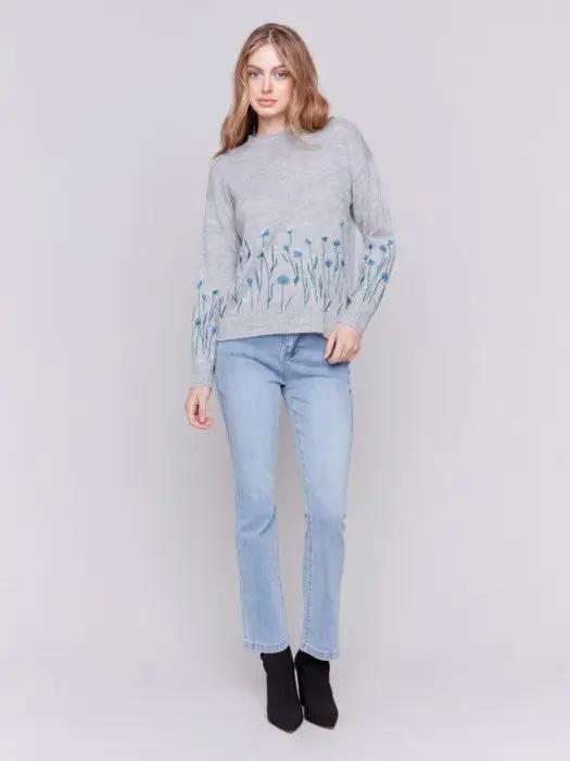 A woman with long hair is wearing the Charlie B Plush Knit Embroidered Flower Sweater featuring gray fabric adorned with blue and green floral details, paired with light blue jeans, creating a look that exudes cozy warmth.