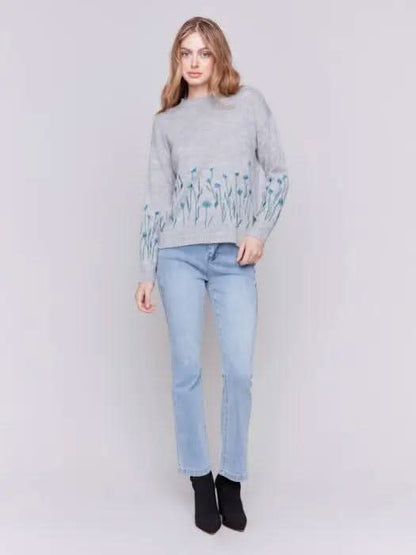 A woman with long hair is wearing the Charlie B Plush Knit Embroidered Flower Sweater featuring gray fabric adorned with blue and green floral details, paired with light blue jeans, creating a look that exudes cozy warmth.