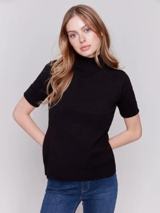 A stylish individual with long hair stands against a plain gray background, donning the Short Sleeve Mock Neck Sweater by Charlie B in black and a pair of comfortably fitting blue jeans.