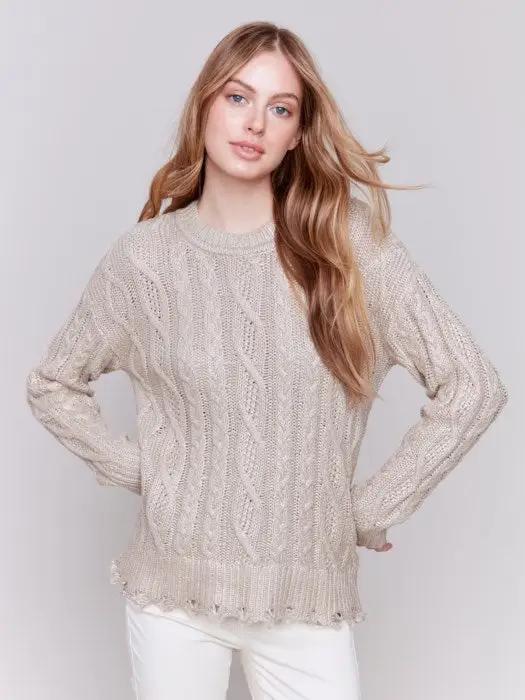 A person with long hair wears a Charlie B Metallic Cable Knit Sweater and white pants, posing against a plain background.