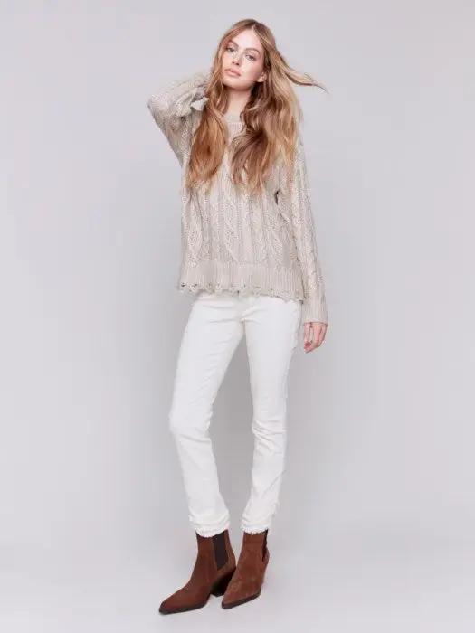 A person with long hair wears a Charlie B Metallic Cable Knit Sweater and white pants, posing against a plain background.