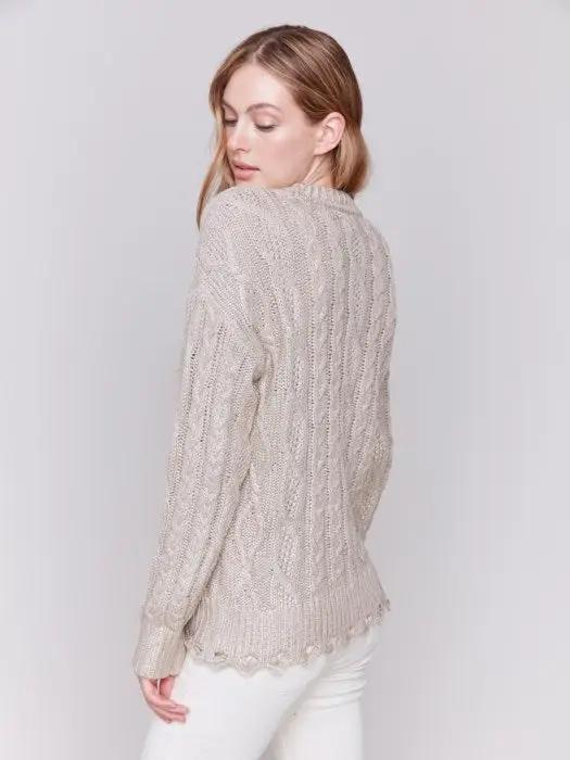 A person with long hair wears a Charlie B Metallic Cable Knit Sweater and white pants, posing against a plain background.