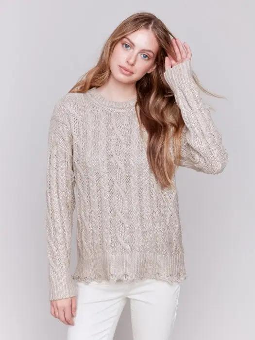 A person with long hair wears a Charlie B Metallic Cable Knit Sweater and white pants, posing against a plain background.