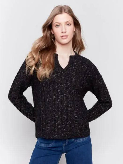 A person with long hair wearing the black Sparkled Yarn Cable Sweater from Charlie B, crafted from ultra-soft material, paired with blue jeans stands against a plain background, looking at the camera.