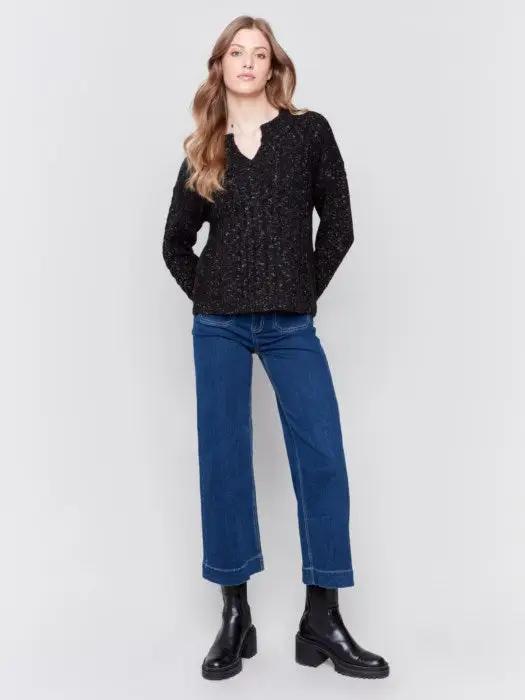 A person with long hair wearing the black Sparkled Yarn Cable Sweater from Charlie B, crafted from ultra-soft material, paired with blue jeans stands against a plain background, looking at the camera.