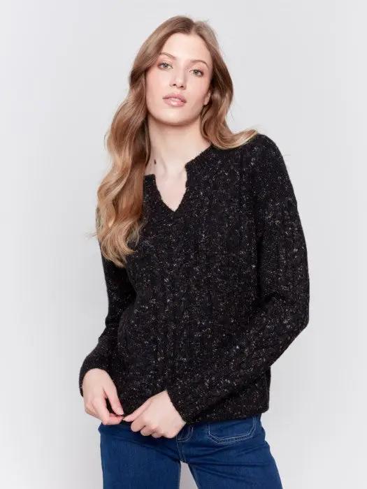 A person with long hair wearing the black Sparkled Yarn Cable Sweater from Charlie B, crafted from ultra-soft material, paired with blue jeans stands against a plain background, looking at the camera.