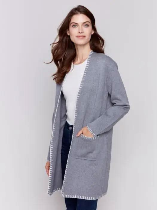 A stylish woman, adorned in a cozy Charlie B Blanket Stitch Cardigan featuring white trim, layered over a white shirt and paired with dark jeans, stands against a plain background.