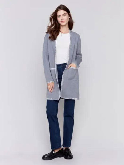 A stylish woman, adorned in a cozy Charlie B Blanket Stitch Cardigan featuring white trim, layered over a white shirt and paired with dark jeans, stands against a plain background.