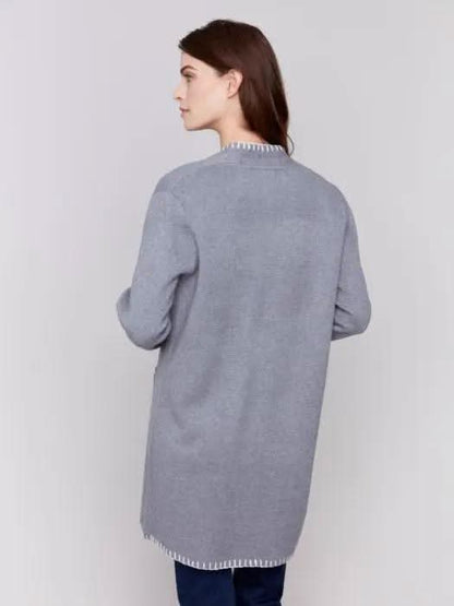 A stylish woman, adorned in a cozy Charlie B Blanket Stitch Cardigan featuring white trim, layered over a white shirt and paired with dark jeans, stands against a plain background.