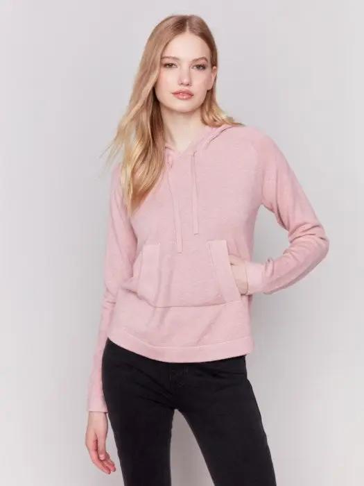 A person with long blonde hair wearing a light pink plush knit hoodie from Charlie B, with hands in the front kangaroo pocket, and black pants, stands against a plain white background.