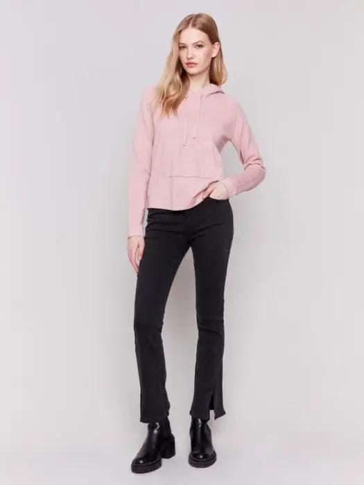 A person with long blonde hair wearing a light pink plush knit hoodie from Charlie B, with hands in the front kangaroo pocket, and black pants, stands against a plain white background.