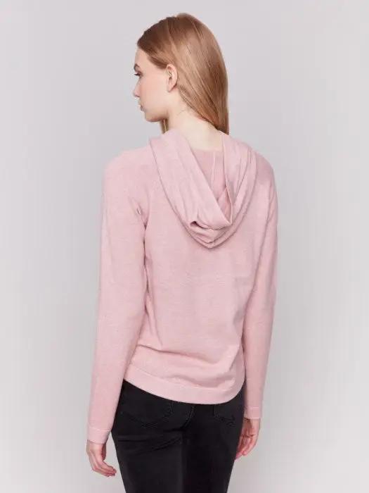 A person with long blonde hair wearing a light pink plush knit hoodie from Charlie B, with hands in the front kangaroo pocket, and black pants, stands against a plain white background.