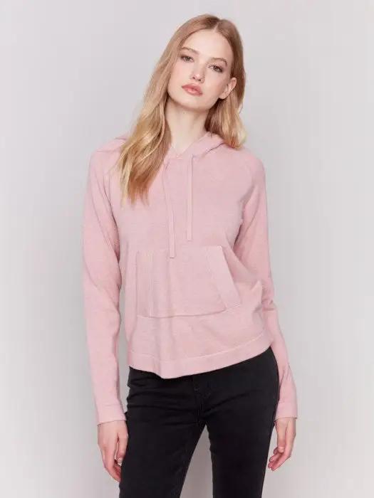 A person with long blonde hair wearing a light pink plush knit hoodie from Charlie B, with hands in the front kangaroo pocket, and black pants, stands against a plain white background.