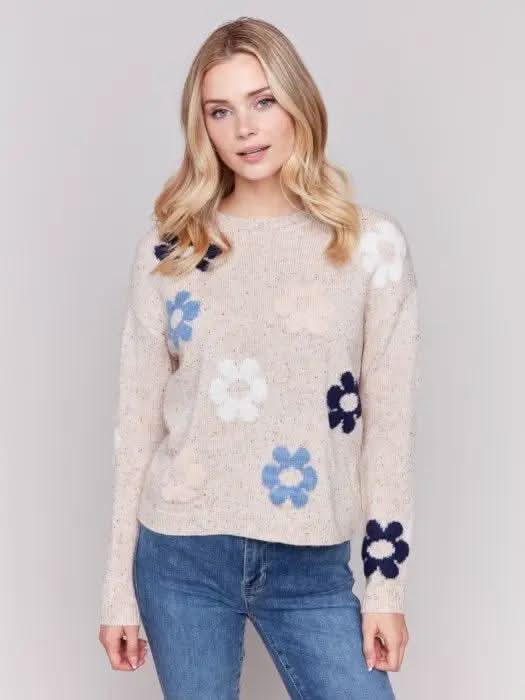 A person with long blonde hair wearing a Charlie B Neps Yarn Knit Sweater adorned with colorful flower patterns and blue jeans stands with hands in pockets against a plain background, perfect for cooler weather.