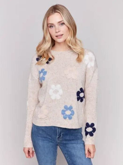 A person with long blonde hair wearing a Charlie B Neps Yarn Knit Sweater adorned with colorful flower patterns and blue jeans stands with hands in pockets against a plain background, perfect for cooler weather.