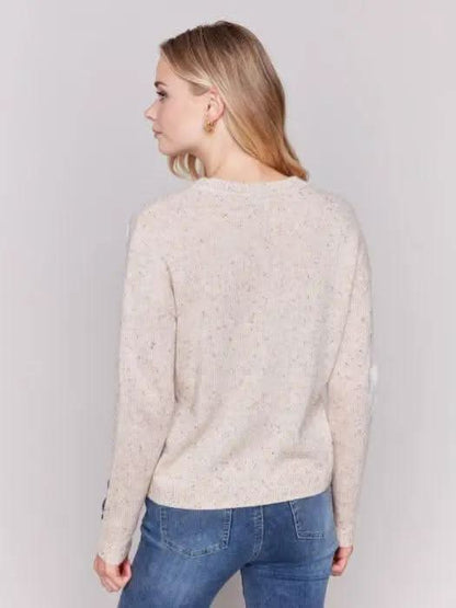 A person with long blonde hair wearing a Charlie B Neps Yarn Knit Sweater adorned with colorful flower patterns and blue jeans stands with hands in pockets against a plain background, perfect for cooler weather.