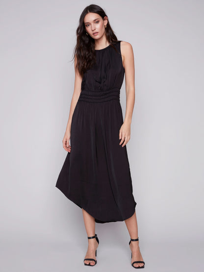 A woman in a Charlie B sleeveless black dress with an elastic smocking waist and strappy heels stands against a plain background.