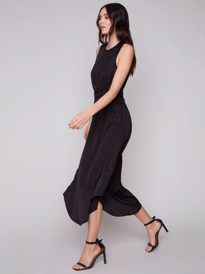A woman in a Charlie B sleeveless black dress with an elastic smocking waist and strappy heels stands against a plain background.