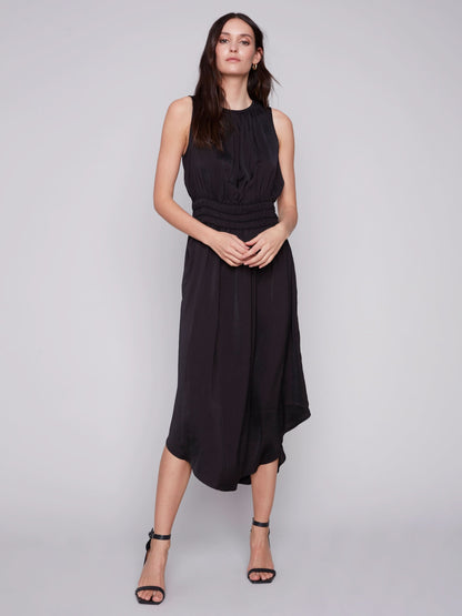 A woman in a Charlie B sleeveless black dress with an elastic smocking waist and strappy heels stands against a plain background.