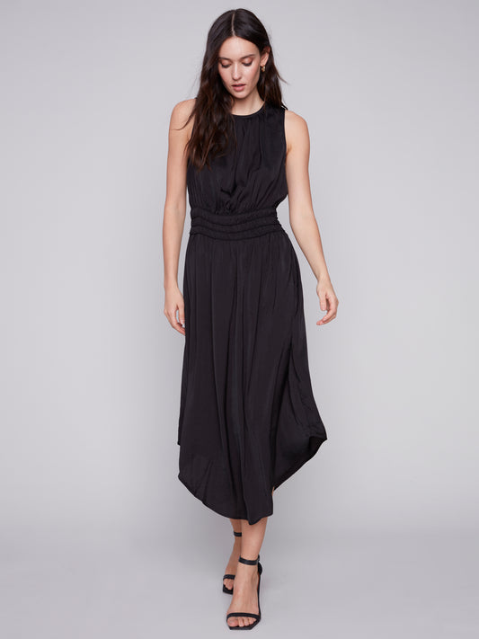 A woman in a Charlie B sleeveless black dress with an elastic smocking waist and strappy heels stands against a plain background.