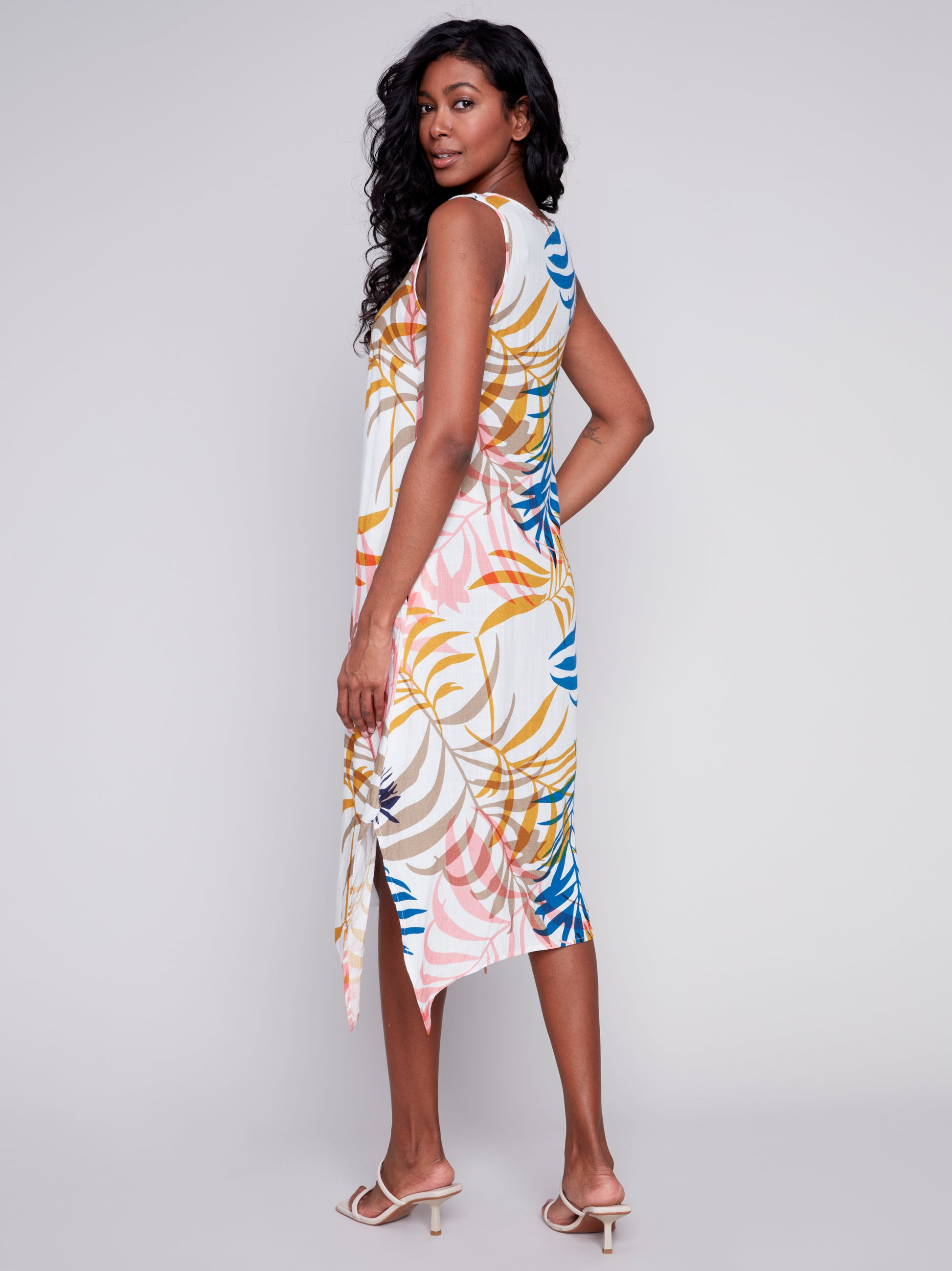 Woman standing in a studio, wearing a colorful tropical print Charlie B midi dress with slits and pockets and beige high heels, smiling at the camera.