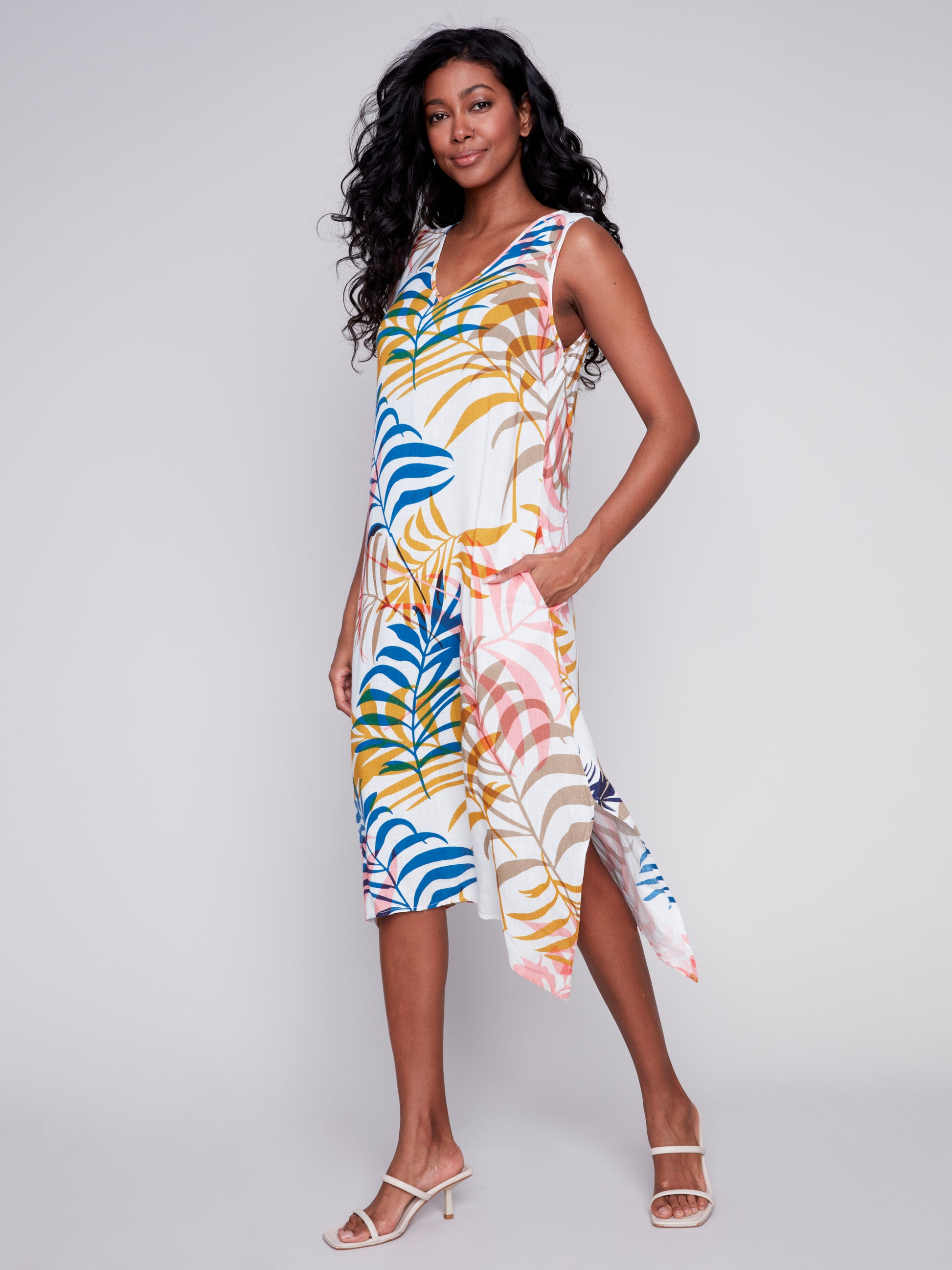 Woman standing in a studio, wearing a colorful tropical print Charlie B midi dress with slits and pockets and beige high heels, smiling at the camera.