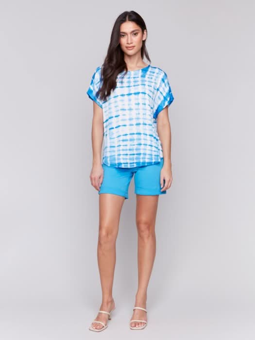 A woman faces forward in a Charlie B Tie Dye Dolman Short Sleeve Relaxed Top, blue and white, paired with blue shorts against a plain light gray background.