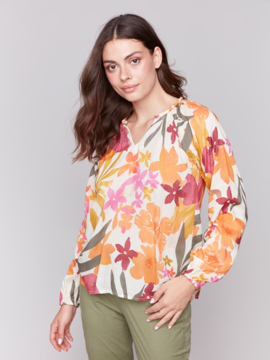 A person with long hair wearing a Charlie B Printed Viscose Long Sleeve Popover Blouse and green pants poses against a plain background, one hand in their pocket.