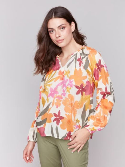 A person with long hair wearing a Charlie B Printed Viscose Long Sleeve Popover Blouse and green pants poses against a plain background, one hand in their pocket.