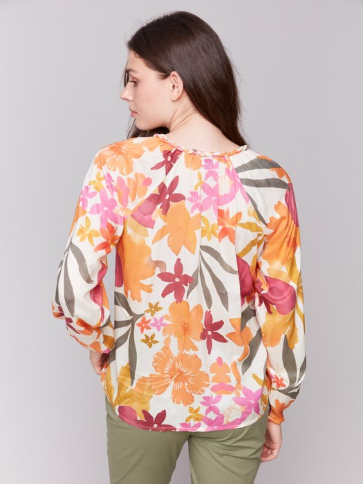 A person with long hair wearing a Charlie B Printed Viscose Long Sleeve Popover Blouse and green pants poses against a plain background, one hand in their pocket.