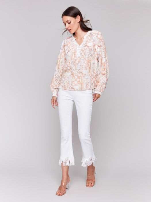 A woman poses with arms crossed in a Charlie B Raglan Blouse with a V-notch neckline and intricate pastel embroidery, paired with white pants, adding elegance to her ensemble.