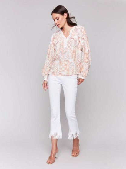 A woman poses with arms crossed in a Charlie B Raglan Blouse with a V-notch neckline and intricate pastel embroidery, paired with white pants, adding elegance to her ensemble.