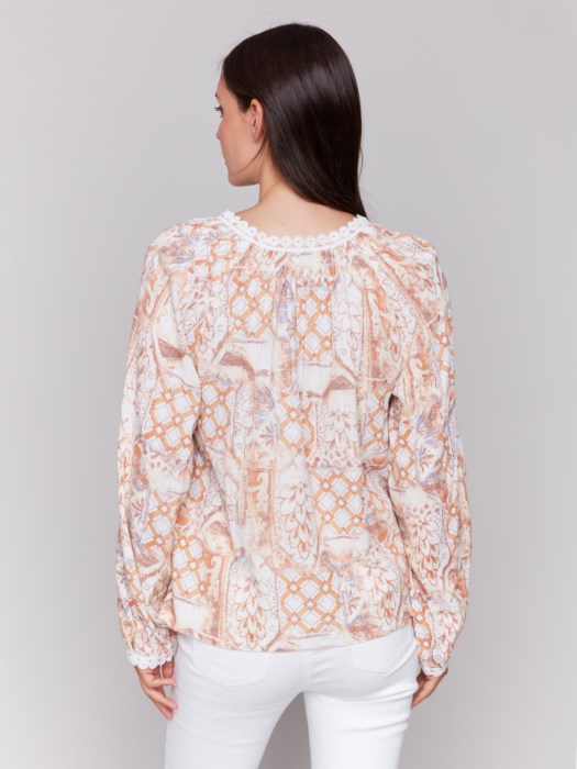 A woman poses with arms crossed in a Charlie B Raglan Blouse with a V-notch neckline and intricate pastel embroidery, paired with white pants, adding elegance to her ensemble.