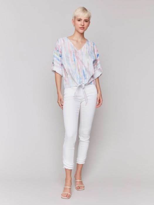 A person stands against a neutral background wearing the Charlie B 3/4 Rolled Cuff Sleeve V Neck in pastel tie-dye, paired with white cropped pants and sandals, one hand in their pocket for a relaxed, casual vibe.