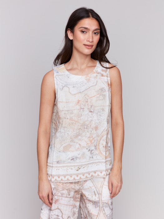 A woman with long hair wears a Charlie B Printed Sleeveless Linen Blend Top and light-patterned pants against a plain gray background, the boat neck top adding elegance to the minimalist setting.