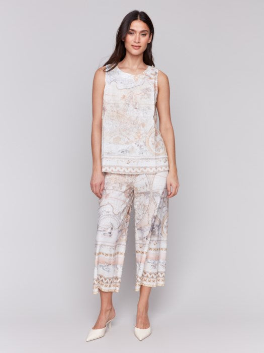 A woman with long hair wears a Charlie B Printed Sleeveless Linen Blend Top and light-patterned pants against a plain gray background, the boat neck top adding elegance to the minimalist setting.