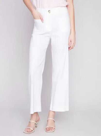 A woman standing against a gray backdrop, wearing a pink sleeveless top and white Charlie B cropped straight leg pants, paired with white heeled sandals.
